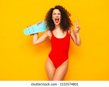 Young beautiful smiling woman posing near yellow wall in studio.Sexy model in red swimwear bathing suit.Positive female with curls hairstyle. Holding penny skateboard.Happy and cheerful