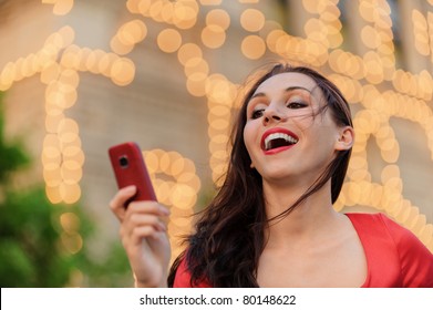 Young Beautiful Smiling Woman In Red Dress Reads Text Message On Mobile Phone Against Evening Illumination.