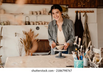 Young beautiful smiling woman sculpting a small clay bowl and looking at camera while standing in a cozy pottery workshop - Powered by Shutterstock