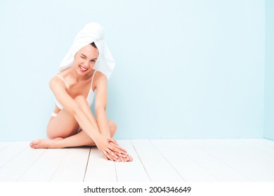 Young beautiful smiling woman in white lingerie.Sexy carefree model in underwear and towel on head posing near light blue wall in studio. Positive and happy female enjoying morning.Sitting and winking