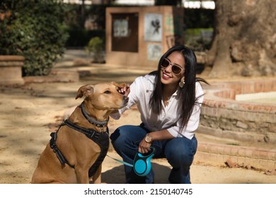 Young Beautiful South American Woman In White Shirt And Sunglasses, Bending Down, Petting Her Dog In A Park. Concept Animals, Dogs, Friends, Breed.