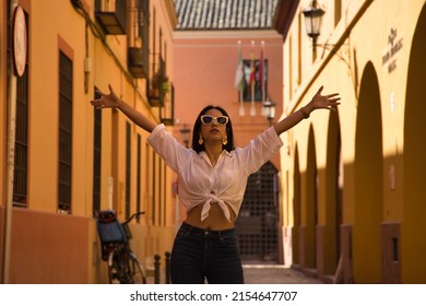 Young And Beautiful South American Woman In White Knotted Shirt, Jeans And Sunglasses With Open Arms In The Middle Of A Cobblestone Street. Concept Beauty, Fashion, Vacation, Travel, Urban, City.