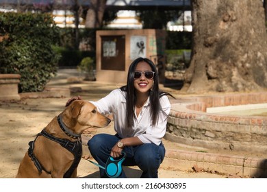 Young Beautiful South American Woman In White Shirt And Sunglasses, Bending Down, Petting Her Dog In A Park. Concept Animals, Dogs, Friends, Breed.