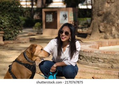 Young Beautiful South American Woman In White Shirt And Sunglasses, Bending Down, Petting Her Dog In A Park. Concept Animals, Dogs, Friends, Breed.