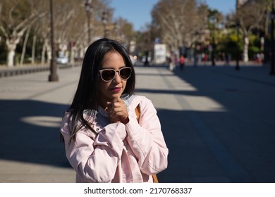 Young beautiful South American woman in a pink jacket and sunglasses, hands clasped praying. Concept religion, prayer, catholic, faith.