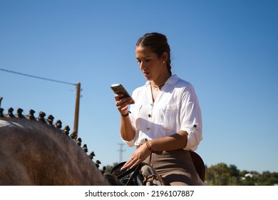 Young And Beautiful Spanish Woman On A White And Grey Horse Consulting Her Mobile Phone. The Woman Is Wearing A Riding Uniform. Thoroughbred And Equine Concept.