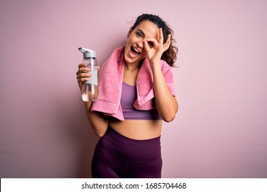 Young beautiful sportswoman with curly hair doing sport drinking bottle of water using towel with happy face smiling doing ok sign with hand on eye looking through fingers