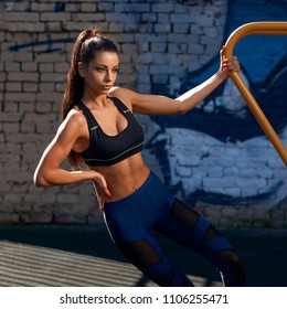 Young beautiful sporty athlete woman with pony tail brunette hair in blue leggings and black top standing at outdoor workout gym. Portrait of sexy girl at sunset