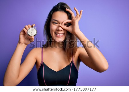Young beautiful sporty girl doing sport using stopwatch over isolated purple background with happy face smiling doing ok sign with hand on eye looking through fingers