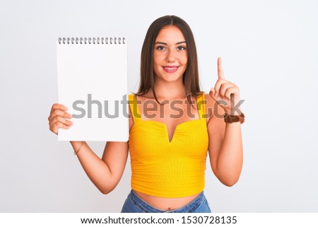Young beautiful student girl holding notebook standing over isolated white background surprised with an idea or question pointing finger with happy face, number one