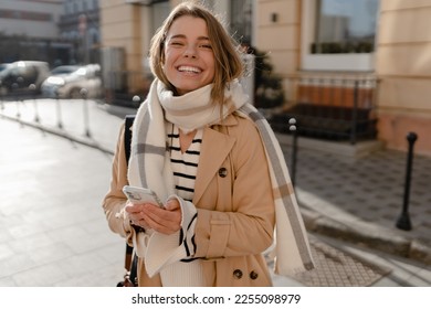 young beautiful stylish woman walking in winter street wearing beige coat, scarf, smiling happy cold season fashion trend, holding using smart phone - Powered by Shutterstock