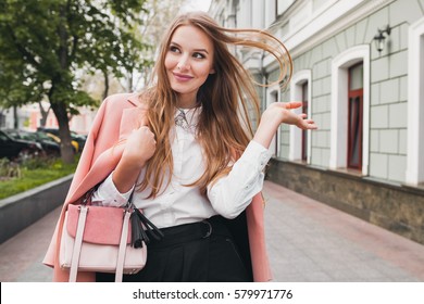 young beautiful stylish woman walking in pink coat, holding purse in hands, smiling, happy, street style, spring summer trend, black skirt, white shirt, flirty