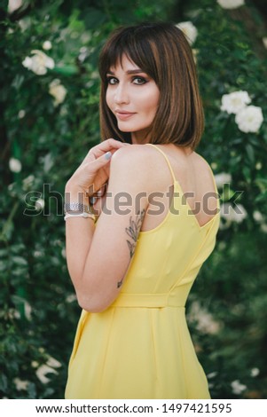 Young beautiful stylish woman in yellow jumpsuit with tattoo on her hand posing near roses flowers in a garden.
