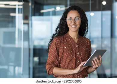Young beautiful and successful business woman with tablet computer and glasses, smiling and looking out the window, Hispanic woman working inside a modern office building at work.