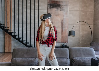 Young Beautiful Swedish Woman In Plaid Shirt And White Pants Using Vr Glasses At Home. Relaxing Swedish Girl In Virtual Reality Headset Standing In Room With Sofa, Stairs, Playing Games. Leisure