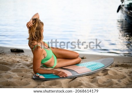 young beautiful tanned woman in swimsuit with wakeboard resting on sandy beach near the water