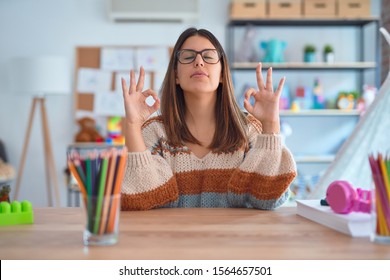 Young Beautiful Teacher Woman Wearing Sweater And Glasses Sitting On Desk At Kindergarten Relaxed And Smiling With Eyes Closed Doing Meditation Gesture With Fingers. Yoga Concept.