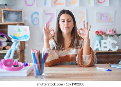 Young Beautiful Teacher Woman Wearing Sweater And Glasses Sitting On Desk At Kindergarten Relax And Smiling With Eyes Closed Doing Meditation Gesture With Fingers. Yoga Concept.