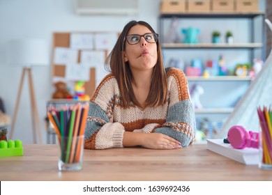 Young Beautiful Teacher Woman Wearing Sweater And Glasses Sitting On Desk At Kindergarten Making Fish Face With Lips, Crazy And Comical Gesture. Funny Expression.