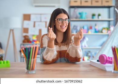 Young Beautiful Teacher Woman Wearing Sweater And Glasses Sitting On Desk At Kindergarten Doing Money Gesture With Hands, Asking For Salary Payment, Millionaire Business