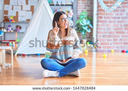 Young beautiful teacher woman wearing sweater and glasses sitting on the floor at kindergarten pointing finger up with successful idea. Exited and happy. Number one.