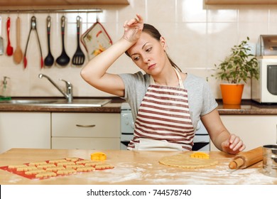 The Young Beautiful Tired Woman 30-35 Years In Apron Sitting At A Table With Dough And Rolling Pin And Going To Prepare A Christmas Ginger Cakes In The Light Kitchen. Cooking Home. Prepare Food.