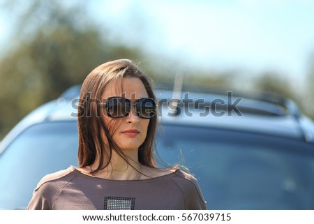 Young beautiful white girl in a brown sports suit stands near a grey and blue car in a field of dandelions. Spring portrait.