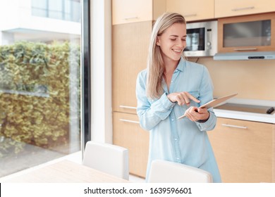 Young Beautiful Wife Uses Smarthome App On Tablet. Electronic Assistant In Household Chores. Modern Gadgets And Digital Devices. Bright Kitchen Interior, Large Windows. Online Store, Booking Hotel