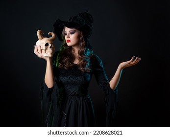 A young beautiful witch in a conical hat holds a devil's skull in her hands and makes a spell with her hand. Halloween Costume Party, Witches' Sabbath