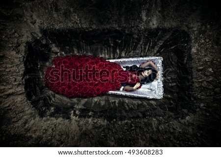 Young beautiful witch in open coffin. Halloween day. White woman in elegant long dress. A lot of red roses of velvet. Big and deep hole in the ground.