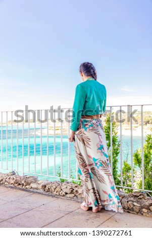 A young and beautiful woman, alone in a luxury hotel, contemplates, thoughtfully, the panorama of the turquoise sea, in Salento, Puglia, Italy. Elegant dress with green jacket. Terrace on the sea.