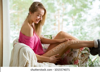 Young beautiful woman applying moisturizing lotion on body skin in armchair. Sitting in pajamas with tube of moisturizer, touching legs. Heart drawn with cream. Love your body and skin care concept.