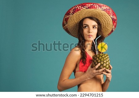 young beautiful woman in bikini and sombrero drinking tropic cocktail looking up left isolated on blue background