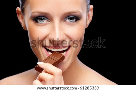 Young beautiful woman bites off slice from chocolate bar, on black background.