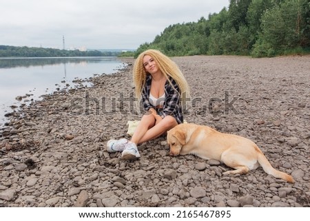 Young beautiful woman with blond curly hair sitting with her labrador retriever dog on the river shore