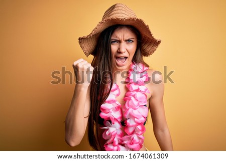 Young beautiful woman with blue eyes on vacation wearing bikini and hawaiian lei angry and mad raising fist frustrated and furious while shouting with anger. Rage and aggressive concept.