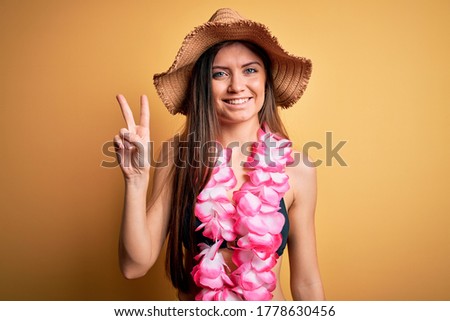 Young beautiful woman with blue eyes on vacation wearing bikini and hawaiian lei showing and pointing up with fingers number two while smiling confident and happy.