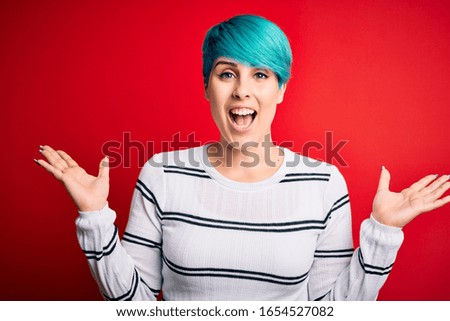 Young beautiful woman with blue fashion hair wearing striped sweater over red background celebrating crazy and amazed for success with arms raised and open eyes screaming excited. Winner concept