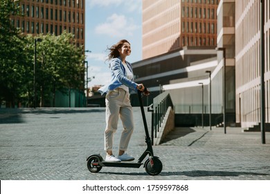 Young beautiful woman in a blue jacket smiles and rides an electric scooter to work along office buildings - Powered by Shutterstock