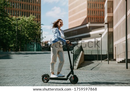 Young beautiful woman in a blue jacket smiles and rides an electric scooter to work along office buildings