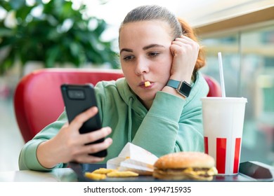 Young Beautiful Woman, Bored Girl Eating Fast Junk Food: Burger, Hamburger, Potato French Fries And Soda At Food Court At Mall And Looking At Her Cell Mobile Phone, Smartphone. Bad Unhealthy Habits 