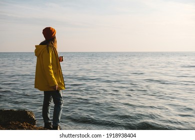 Young beautiful woman in bright yellow coat stands on the seashore or ocean with cup of tea, coffee and enjoy the view. Concept of freedom, thoughtfulness, mindfulness.