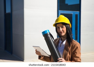 Young Beautiful Woman In Brown Coat, Yellow Workman's Helmet And Blueprint Tube, Working With Her Tablet. Business Concept, Architect, Construction, Working Woman, Empowerment.