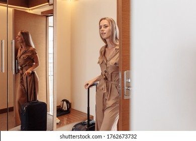 Young Beautiful Woman In Brown Overalls With Suitcase Settles In Cool Hotel Room While Travelling. Charming Successful Happy Female Model Moves To New Apartment And Enjoys Changes. Girl Is Checked In