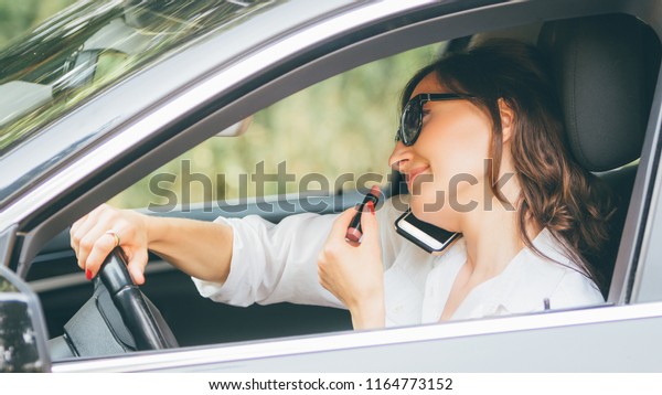 Young beautiful woman in a car talk on the\
phone and dye her lips while driving - multitasking lady drives\
with lipstick and\
smartphone