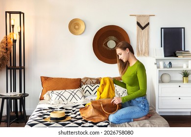 Junge schöne Frau in ungezwungenen Kleidern, die zu Hause auf dem Bett sitzt und ihren gelben Herbstschal in die Lederhandtasche steckt, einen Spaziergang im Freien macht, in einem stilvoll eingerichteten und gemütlichen Zimmer verbringt