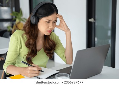 Young, beautiful woman in casual clothes feeling stressed as she sits at a desk in her dedicated workspace delves into online study or diligently works on her project with her laptop, notebook,and hea - Shutterstock ID 2366987677
