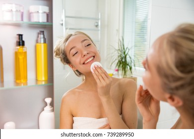 Young Beautiful Woman Cleaning Her Face Skin With Cotton Pad In Bathroom. Standing In Towel, Looking In The Mirror, Laughing And Having Fun. Morning Skincare Routine.  