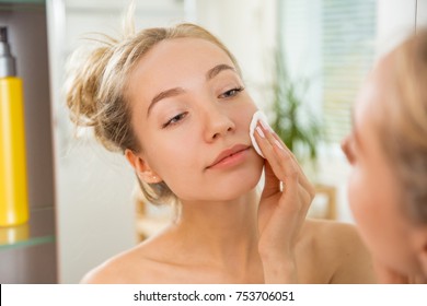 Young beautiful woman cleaning her face skin with cotton pad in bathroom. Standing in towel, looking in the mirror, laughing and having fun. Morning skincare routine.   - Powered by Shutterstock