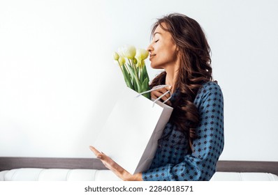 Young beautiful woman closed her eyes and smells bouquet of tulip flowers with pleasure. Female shopper in casual clothing holding two white paper shopping bags. White background. Copy space.